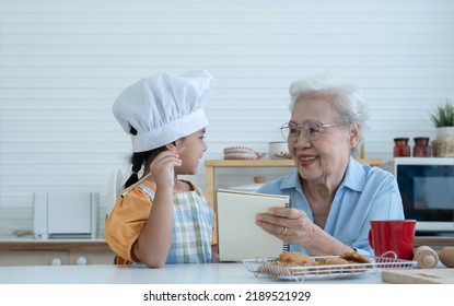 Asian Family Grandmother And Grandchild Have Fun Cooking At Home Kitchen Together, Little Cute Granddaughter With Chef Hat And Apron Is Writing Down Recipe And How To Make Cookie From Grandma