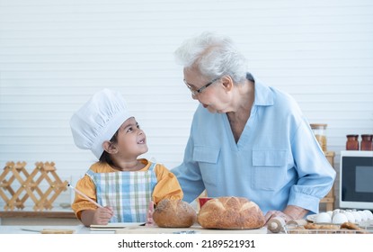 Asian Family Grandmother And Grandchild Have Fun Cooking At Home Kitchen Together, Little Cute Granddaughter With Chef Hat And Apron Is Writing Down Recipe And How To Make Bread Or Cookie From Grandma