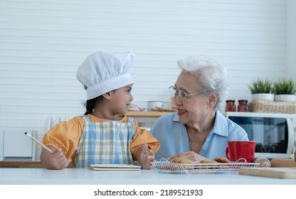 Asian Family Grandmother And Grandchild Have Fun Cooking At Home Kitchen Together, Little Cute Granddaughter With Chef Hat And Apron Is Writing Down Recipe And How To Make Bread Or Cookie From Grandma