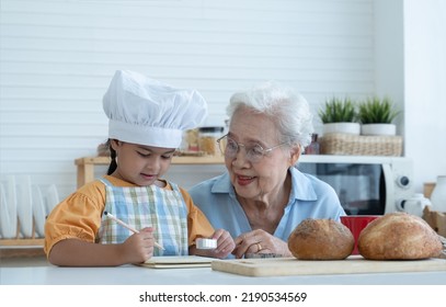 Asian Family Grandmother And Grandchild Have Fun Cooking At Home Kitchen Together, Little Cute Granddaughter With Chef Hat And Apron Is Writing Down Recipe And How To Make Bread Or Cookie From Grandma