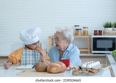 Asian Family Grandmother And Grandchild Have Fun Cooking And Baking Cookies At Home Kitchen Together, Little Cute Granddaughter With Chef Hat And Apron Is Eating Homemade Cookie From Grandma