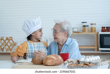 Asian Family Grandmother And Grandchild Have Fun Cooking And Baking Cookies At Home Kitchen Together, Little Cute Granddaughter With Chef Hat And Apron Is Eating Homemade Cookie From Grandma