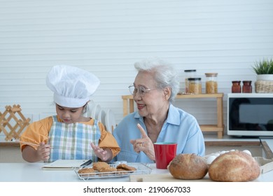 Asian Family Grandmother And Grandchild Have Fun Cooking At Home Kitchen Together, Little Cute Granddaughter With Chef Hat And Apron Is Writing Down Recipe And How To Make Bread Or Cookie From Grandma