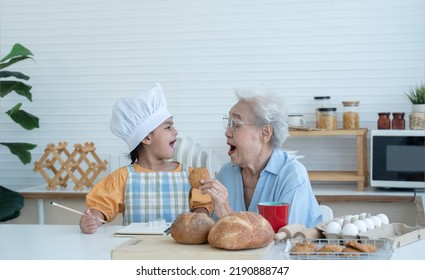 Asian Family Grandmother And Grandchild Have Fun Cooking And Baking Cookies At Home Kitchen Together, Little Cute Granddaughter With Chef Hat And Apron Is Eating Homemade Cookie From Grandma