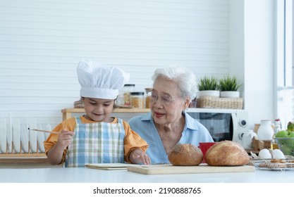 Asian Family Grandmother And Grandchild Have Fun Cooking At Home Kitchen Together, Little Cute Granddaughter With Chef Hat And Apron Is Writing Down Recipe And How To Make Bread Or Cookie From Grandma