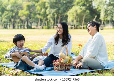 Asian Family Having Picnic In Fresh Green Park Together. Mother Touching To Kid With Smile While Grandmother Looking At Child Playing Ukelele Or Mini Guitar With Happiness. Relax Family Activity.