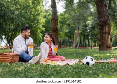 Asian Family Holding Orange Juice Near Food And Football On Grass In Park