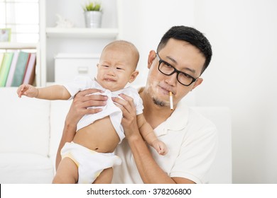 Asian Family At Home. Bad Father Smoking While Holding Baby Together. Cigarette With Lit And Smoke. Unhealthy Lifestyle Or Stop Smoking Concept Photo.