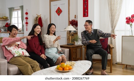 Asian Family Laughing And Pointing With Finger Enjoying Watching Tv Together In Living Room On Chinese Lunar New Year Holiday. Word At Background Translation: Spring And Congratulation