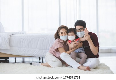 Asian Family In Medical Masks On The Face Looks At The Camera While Standing In The Living Room At Home To Prevent PM2.5 Dust, Smog, Air Pollution And COVID-19. Healthcare Concept.