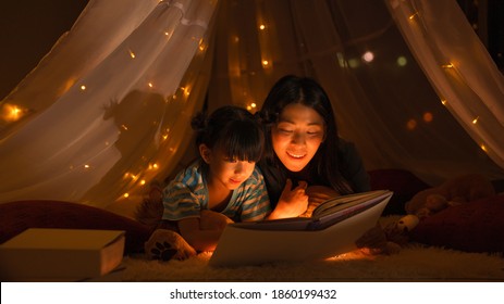 Asian Family Mom And Girl Happy At Night Time In Bedroom At Home Before Bed Time Reading Fantasy Bedtime Story Book Together In Child Tent With Dim Yellow Light
