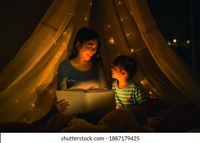Asian family mom and girl happy at night time in bedroom at home before bed time reading fantasy bedtime story book together in kid tent with dim yellow light