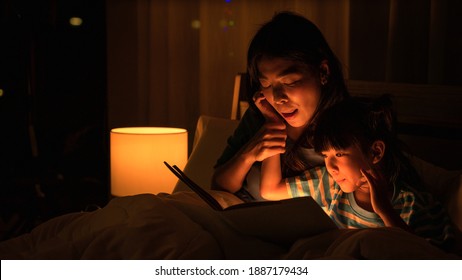 Asian Family Mom And Girl Happy At Night Time In Bedroom At Home Before Bed Time Reading Fantasy Bedtime Story Book Together On Bed With Dim Yellow Light