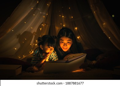 Asian Family Mom And Girl Happy At Night Time In Bedroom At Home Before Bed Time Reading Fantasy Bedtime Story Book Together In Kid Tent With Dim Yellow Light