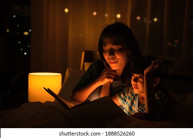 Asian Family Mom And Girl Happy At Night Time In Bedroom At Home Before Bed Time Reading Fantasy Bedtime Story Book Together On Bed With Dim Yellow Light