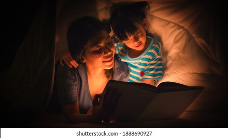 Asian Family Mom And Girl Happy At Night Time In Bedroom At Home Before Bed Time Reading Fantasy Bedtime Story Book Together On Bed Under Blanket With Dim Yellow Light