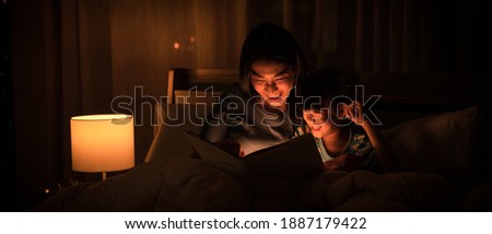 Asian family mom and girl happy at night time in bedroom at home before bed time reading fantasy bedtime story book together on bed with dim yellow light