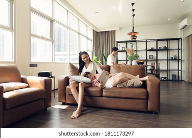 Asian Family Mother Father And Daughter Spending Time Together In The Living Room. Lifestyle Family Relaxing Time In Apartment While The Man Working At Behind With Laptop And Woman And Her Tablet.  