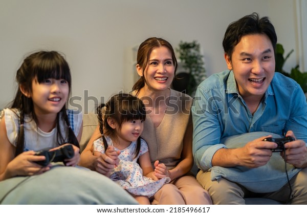 Asian family play video game together in living\
room at home at night. Attractive parent and dauthers sit and\
holding game controller feeling excited and fun with smiling face\
in living room at house.