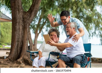 Asian Family Senior Retired Couple With Adult Son Using Digital Tablet Video Call With Their Family Friend On The Beach In Summer Day. Happy Family Enjoy And Having Fun Together In Holiday Vacation