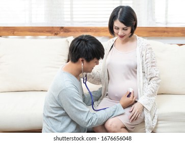 Asian Family Sit Together At Home With Smile.Happy Father Listening Baby Heart Pulse On Mother Tummy By Stethoscope.Dad Feeling Excited With Future Coming Newborn. Family And Mother’s Day Relationship