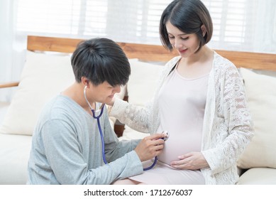 Asian Family Sit Together At Home With Smile.Happy Father Listening Baby Heart Pulse On Mother Tummy By Stethoscope.Dad Feeling Excited With Future Coming Newborn. Family And Mother’s Day Relationship