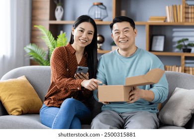 Asian family sitting on sofa unboxing package, smiling and using phone. Happy couple enjoying online shopping and home delivery experience. Cozy home environment with positive emotions - Powered by Shutterstock