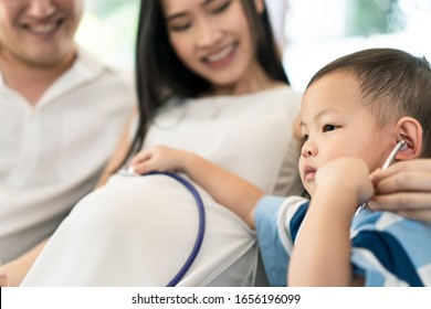 Asian Family Sitting Together At Home With Smile. Young Child Listening Baby Heart Pulse On Mother Tummy By Stethoscope With Father. Kid Feeling Excited With Future Coming Newborn. Family Relationship