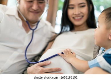 Asian Family Sitting Together At Home With Smile. Young Child Listening Baby Heart Pulse On Mother Tummy By Stethoscope With Father. Kid Feeling Excited With Future Coming Newborn. Family Relationship