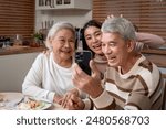 Asian family taking a selfies while having dinner on evening in house. Senior grandparent and daughter using phone take a photo while eating foods celebrate weekend reunion together at table at home.