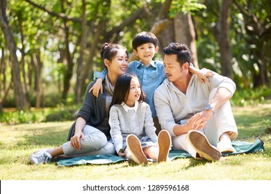 Asian Family With Two Children Having Fun Sitting On Grass Talking Chatting Outdoors In Park