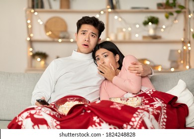 Asian Family Watching Television And Scary Movies On TV Spending Christmas Eve Sitting On Couch In Living Room At Home, Covered With Blanket. Couple Watches Horror During Xmas And New Year Holidays