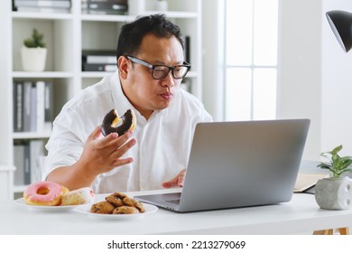 Asian Fat Man Eating Donut, Sweet, Junk Food During Working With Computer Laptop, Unhealthy Eating Concept