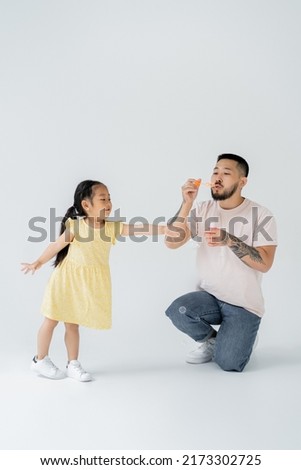 asian father blowing soap bubbles near cheerful preschooler daughter on grey