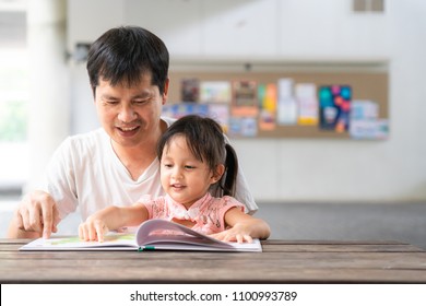 Asian Father And Daughter Are Reading The Storybook Together And They Are Smiling With Happy Moment, Concept Of Learning Activity For Kid In Family Lifestyle. And Role Of Parent Support To The Child.