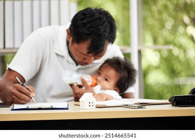 Asian father feeds his baby while writing at desk, showcasing multitasking in work-from-home setting with parenting duties - Powered by Shutterstock