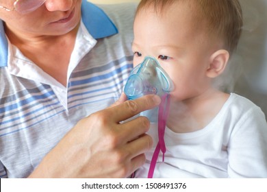 Asian Father Helping His Toddler Son With Inhalation Therapy By The Mask Of Inhaler. Sick Little Kid With Respiratory Problem With Oxygen Mask Breathes Through Nebulizer At Doctor Clinic / Hospital