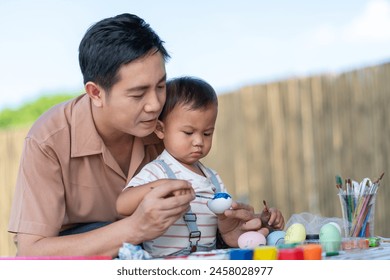 Asian father and his young son engaged in painting Easter eggs with watercolors, sitting on table indoor at home. - Powered by Shutterstock