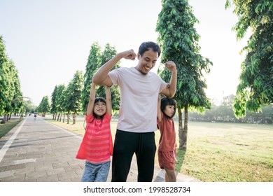 Asian Father And Little Daughter Do Exercises In Outdoor. Healthy Lifestyle Of Family With Child. Strong Dad With Kids Hanging On His Arm