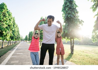 Asian Father And Little Daughter Do Exercises In Outdoor. Healthy Lifestyle Of Family With Child. Strong Dad With Kids Hanging On His Arm