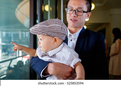 Asian Father And Little Son Dressed In Formal Wear Indoor,baby Wearing Hat And Bow Tie, A 11 Months Toddler Boy With Interest Points A Finger And Looking Outside The Windows,Happy Dad Holding Baby Boy