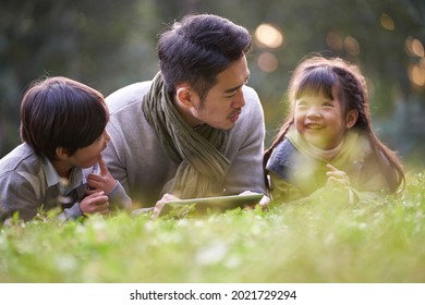 Asian Father Lying On Front On Grass Telling Story To Two Children Happy And Smiling