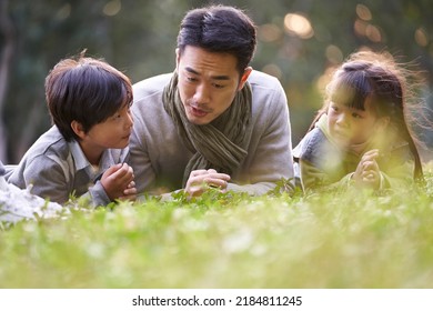 Asian Father Lying On Front On Grass Telling Story To Two Children Happy And Smiling