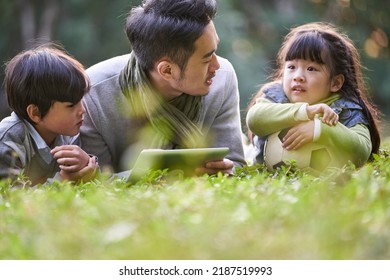 Asian Father Lying On Front On Grass Telling Story To Two Children Happy And Smiling