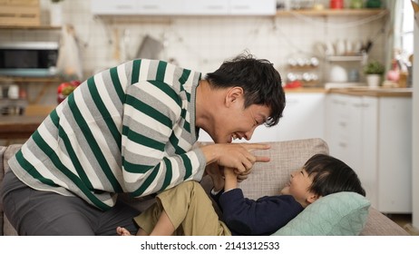 Asian Father Making Silly Face To His Baby Boy. The Child Is Having Fun Playing With His Funny Dad Lying On Living Room Couch At Home