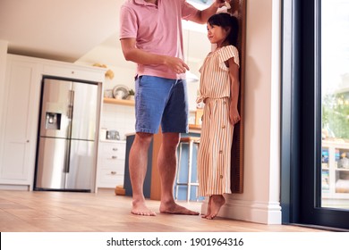 Asian Father Measuring Daughter On Wall Scale At Home As She Stands On Tip Toes
