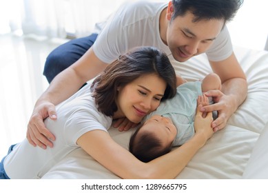 Asian Father And Mother Are Hugging New Newborn Baby On Sofa. They Are Smiling And Warm Touching To The Baby With Love. Father Holding Both Of Baby Hand And Mother Shoulder. Happy Family Concept.