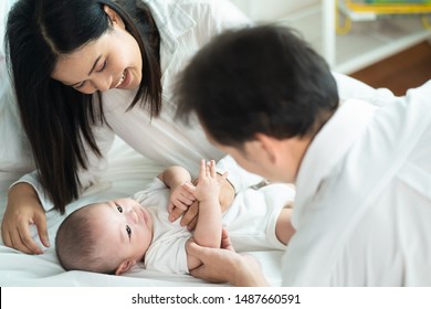 Asian Father And Mother Are Playing With New Newborn Baby On Sofa. They Are Smiling And Warm Touching To The Baby With Love. Parents Holding Both Of Baby Hand. Happy Family Concept.