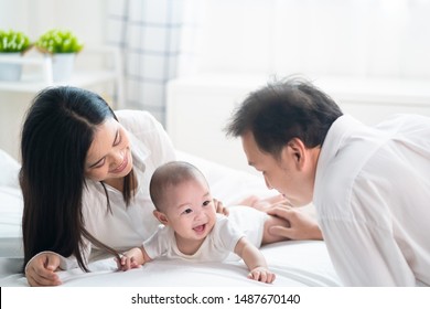 Asian Father And Mother Are Playing With New Newborn Baby On Bed. They Are Smiling And Warm Touching To The Baby With Love. Parents Hugging Baby That Crawling On The Bed. Happy Family Concept.