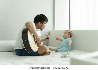 Asian Father playing acoustic guitar for baby in blue clothes on bed, baby reaching out in excitement, morning light through window - Powered by Shutterstock